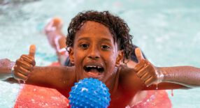 jongen ligt op foamboard in het water met waterspeelgoed en steekt lachend twee duimen in de lucht