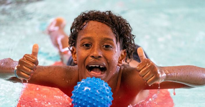 jongen ligt op foamboard in het water met waterspeelgoed en steekt lachend twee duimen in de lucht