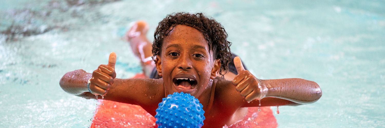 jongen ligt op foamboard in het water met waterspeelgoed en steekt lachend twee duimen in de lucht